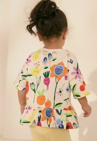 Kortærmet bluse med blomstermotiv og flæsedetaljer, prydet med farverige blomster og grøntsager på en lys baggrund, lavet af blød stof.
