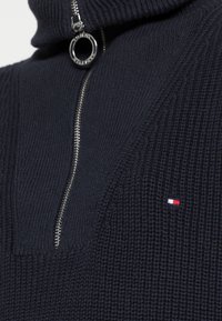 Pull en tricot marine avec col montant et fermeture éclair. Présente un petit logo brodé en rouge, blanc et bleu sur la poitrine.