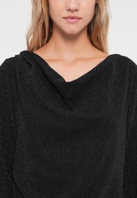 Čierny štruktúrovaný top s drapovaným výstrihom a jemným vertikálnym ryhovaním. Materiál má mierny lesk, čo zvyšuje jeho vizuálnu hĺbku.