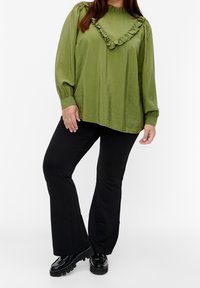 Kvinna iklädd en olivgrön volangblus, svarta utsvängda byxor och svarta klumpiga loafers, stående mot en vit bakgrund.