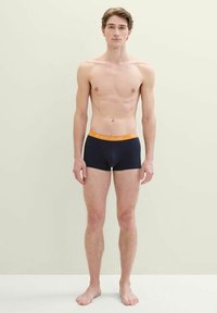 Férfi sötétkék boxer alsónadrág narancssárga derékkal, amelyen a "TOM TAILOR" logó látható. Puha anyagból készült. A modell mezítláb áll egy világos háttér előtt.