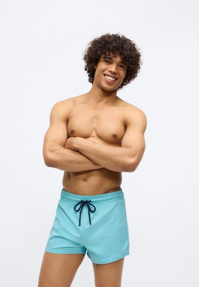 Smejošs jauns vīrietis ar čirkainiem matiem stāv kaila auguma, ar rokām saliktām krūtīs, ģērbies gaiši zilās peldbiksēs pret vienkāršu fona.