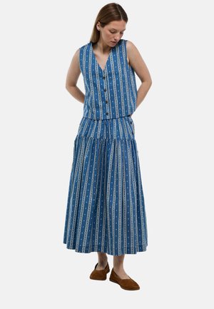 Femme portant un haut bleu sans manches boutonné et une longue jupe assortie à rayures verticales blanches et points, chaussures plates marron, debout les mains dans le dos.