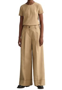 Beige t-shirt med kort ärm i kombination med vida, högmidjade byxor i matchande beige, med synlig sömnad och knappstängning.