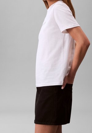 Personne portant un t-shirt blanc uni à manches courtes et un short noir, debout de profil avec une main dans la poche arrière sur un fond uni.