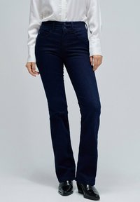 Mujer vestida con jeans de tiro alto en azul oscuro, camisa blanca de manga larga y botas negras de tacón, de pie contra un fondo claro y liso.