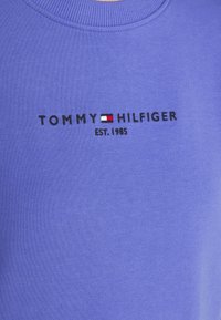 Fioletowy sweter z okrągłym dekoltem, z logo "TOMMY HILFIGER" w kolorze czarnym oraz małym detalem w czerwono-białej flagi. Materiał ma gładką teksturę.