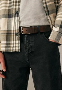 Chemise à carreaux beige, verte et noire sur un t-shirt gris, associée à un jean foncé et une ceinture en cuir marron avec une boucle en argent.