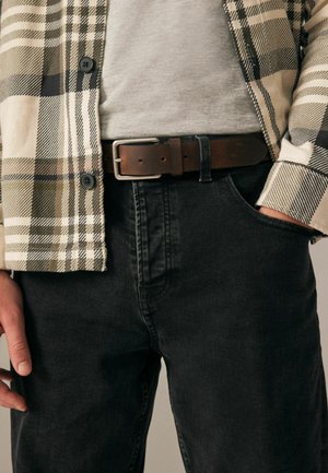 Chemise à carreaux beige, verte et noire sur un t-shirt gris, associée à un jean foncé et une ceinture en cuir marron avec une boucle en argent.