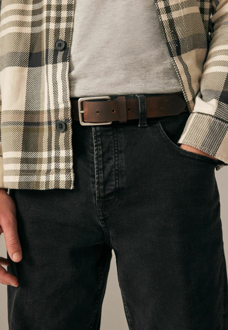 Chemise à carreaux beige, verte et noire sur un t-shirt gris, associée à un jean foncé et une ceinture en cuir marron avec une boucle en argent.