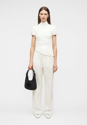 Femme portant un haut blanc à manches courtes côtelé et un pantalon large, tenant un sac à main noir texturé, debout devant un fond uni.