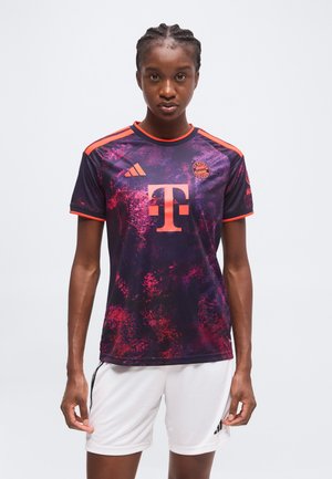 Vrouw met gevlochten haar die een donkerpaarse en roze Bayern München voetbalshirt en witte short draagt, staand tegen een effen achtergrond.