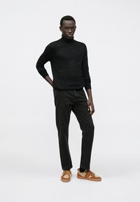 Maglione a collo alto nero abbinato a jeans verde scuro e sneakers beige, con dettagli testurizzati e una vestibilità casual. Posizione in piedi.