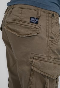 Bruine cargoshorts met meerdere zakken, gestikte naden en een stoffen label. Het materiaal heeft een gladde, matte textuur.