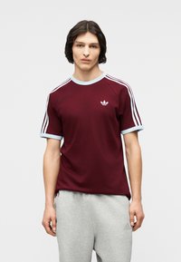 Νεαρός άνδρας που φοράει ένα μπορντό μπλουζάκι Adidas με λευκές ρίγες στα μανίκια και γκρι φόρμα, μπροστά σε ένα απλό ανοιχτό φόντο.