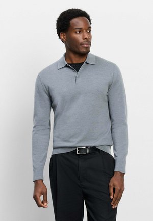 Uomo che indossa una polo grigio chiaro a maniche lunghe sopra una maglia nera con pantaloni neri e cintura nera, che guarda a destra.