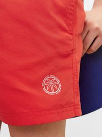 Pantaloni da bagno rossi realizzati in materiale leggero, con un logo circolare bianco che presenta un design di palma vicino all'orlo.