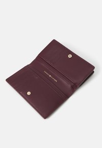 Bordo usnjeni denarnica z zapiranjem na gumb, notranjimi režami za kartice in embosiranim logotipom "TOMMY HILFIGER" v zlatem. Gladka tekstura, minimalističen dizajn.