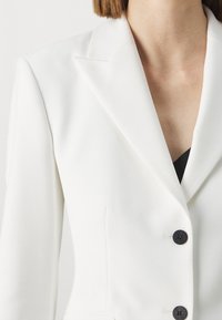 Bílý přizpůsobený blazer s texturovanou tkaninou, klopovými límci a černými knoflíky. Má přiléhavý siluetu a dvojité přední kapsy.