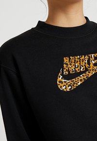 Svart sweatshirt med hög krage, prydd med en leopardmönstrad Nike-logotyp i orange och svart framtill. Mjuk tygtextur.