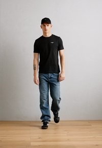 Tricou negru cu guler rotund și logo mic, blugi albaștri și adidași negri. Modelul stă pe un fundal neutru cu podea din lemn.