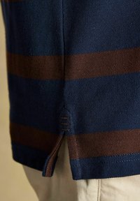 Tuvum tuvu ar jūras zilu un brūnu svītru adījumu ar sāna šķirbu un šuvju detaļām, uzvilkts pār beigu biksēm.