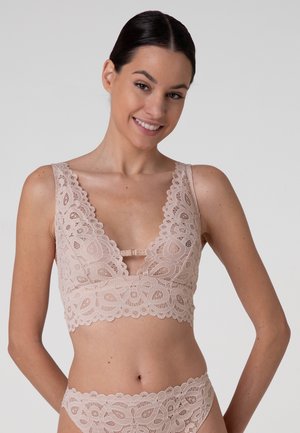 Donna con capelli scuri in un bralette di pizzo rosa e mutandine abbinate, che sorride leggermente su uno sfondo chiaro uniforme.