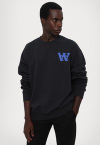 Svart hettegenser med en blå grafisk "W" logo på brystet, ribbede mansjetter og rund halsdesign. Stoffet virker mykt og strukturert.