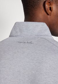 Grå polo skjorte med høy krage, laget av et mykt materiale med en subtil tekstur. Merkevaren sin logo er brodert på baksiden.