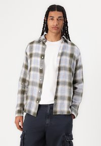 Camisa de franela a cuadros en verde y azul con un diseño de botones, que presenta un corte holgado, manga larga y botones blancos. Llevada sobre una camiseta blanca.