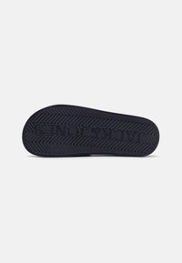 Semelle de chaussure en caoutchouc noir avec un motif en zigzag texturé, avec un logo en relief et un talon arrondi. Fond uni.