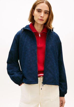 Nő sötétkék mintás cipzáras dzsekiben, piros félig cipzáras pulóver fölött és krémszínű nadrágban, zsebre tett kézzel, egyszínű háttér előtt állva.