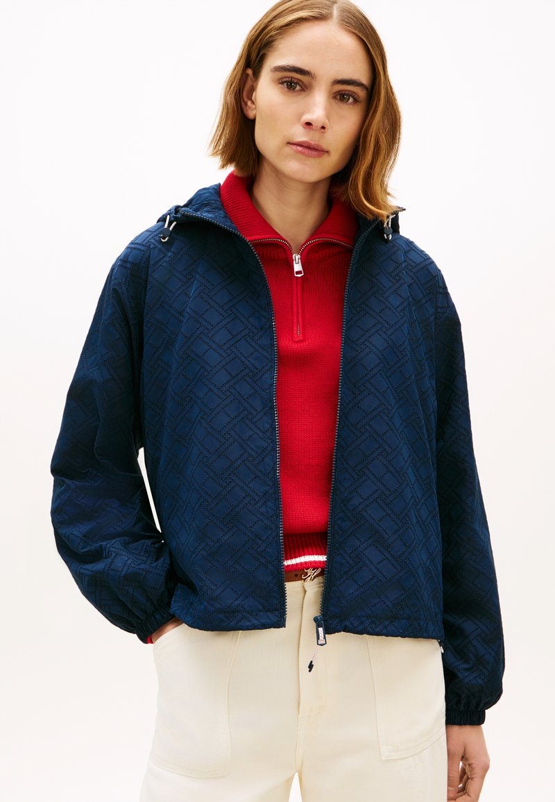 Γυναίκα με ναυτικό μάλλινο τζάκετ με φερμουάρ και σχέδιο, από πάνω από κόκκινο πουλόβερ μισό φερμουάρ και κρεμ παντελόνι, στέκεται με τα χέρια στις τσέπες μπροστά σε απλό φόντο.