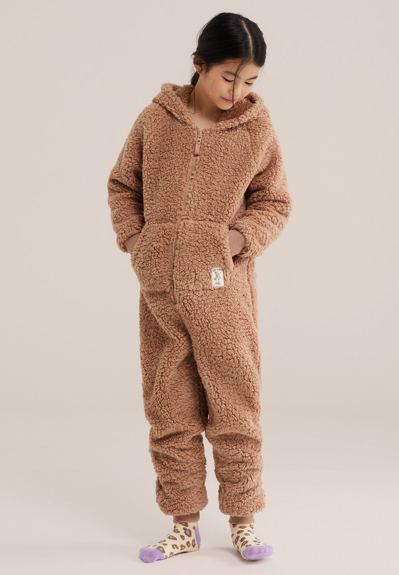 Enfant portant une combinaison à capuche marron et duveteuse, les mains dans les poches et regardant vers le bas, debout sur un fond neutre uni.