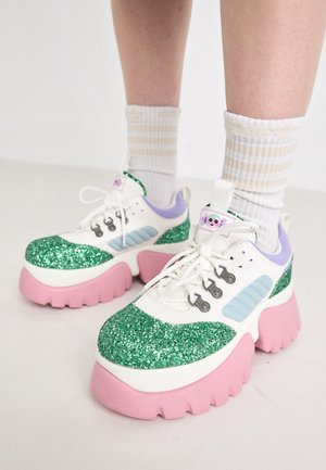 Baskets épaisses avec des bouts pailletés verts, des dessus blancs et bleu pastel, des semelles épaisses roses, et des chaussettes blanches à rayures beige sur les jambes.