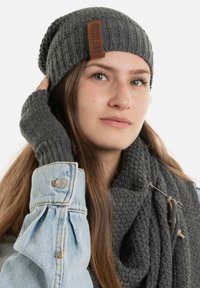 Grijze gebreide beanie met een gestructureerd patroon en leren label. Bijpassende grijze vingerloze handschoenen en een dikke, patroon sjaal. Mouw van een spijkerjack zichtbaar.
