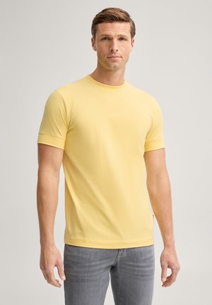 Man draagt een effen geel T-shirt met korte mouwen en grijze jeans, staand tegen een neutrale achtergrond.