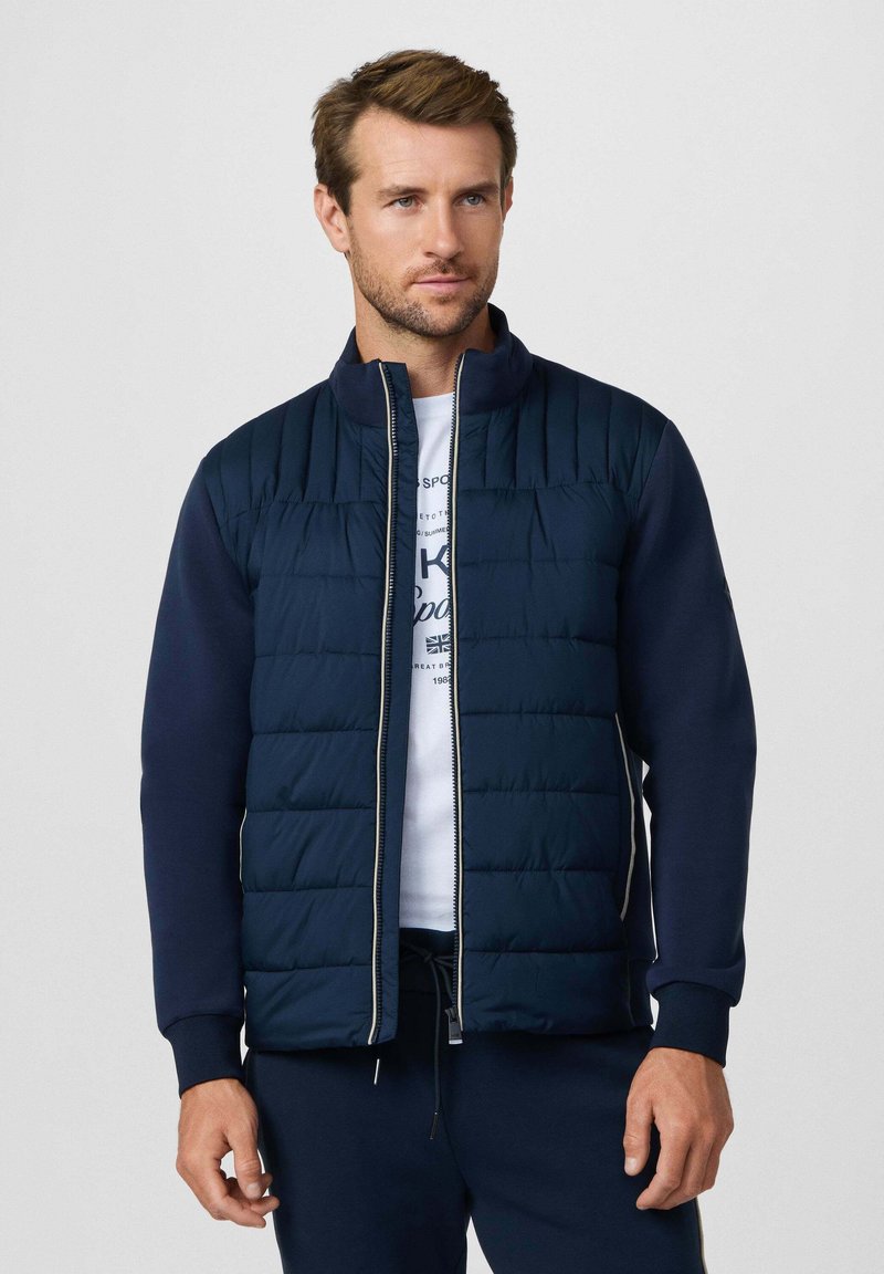 Man in een marineblauwe doorgestikte jas met rits over een wit grafisch t-shirt en bijpassende marineblauwe broek, staand tegen een effen achtergrond.