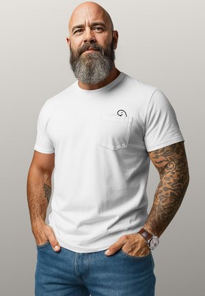 Bearded man with tattooed arms wearing a white t-shirt with a small black design on the pocket and blue jeans, standing with hands in pockets.