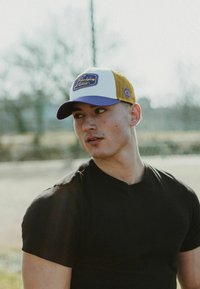 Fehér és sárga hálós dizájnnal rendelkező baseball sapka, lila karimával és téglalap alakú logóval; egy testhezálló fekete pólóval viselve.