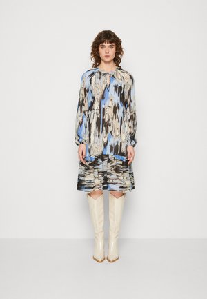 Γυναίκα που στέκεται μπροστά σε έναν απλό τοίχο, φορώντας ένα φόρεμα με μπλε, μπεζ και μαύρο αφηρημένο σχέδιο, με μακριά μανίκια και λευκές μπότες μέχρι το γόνατο.