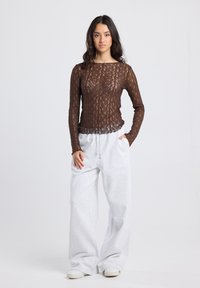 Vrouw staat en draagt een doorzichtige lange mouw bruin kanten top, losse lichtgrijze sweatpants en witte sneakers tegen een effen achtergrond.