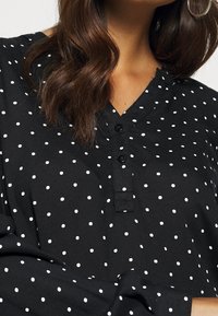 Femme portant un blouse noire à pois blancs, décolleté en V boutonné, et de grandes boucles d'oreilles, les cheveux recouvrant partiellement une épaule.