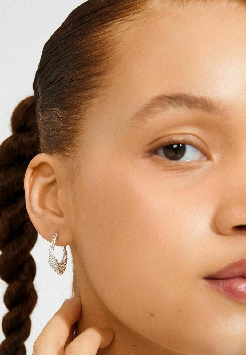 Kvinde med fletning iført sølvfarvede, strukturerede hoops i ørerne, som berører sin hals, viser glat hud og naturlig makeup på lys baggrund.