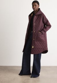 Gequilted Bordeaux-jack met capuchon, voorzien van gouden hardware, twee zijzakken en een licht gebogen zoom, gecombineerd met donkere denimbroek.