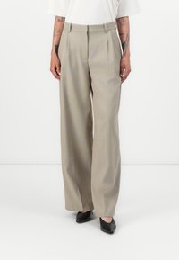 Beige wijde broek met plooien, gemaakt van een gladde stof. Draagt een witte top en zwarte puntige schoenen, met een getailleerde pasvorm.
