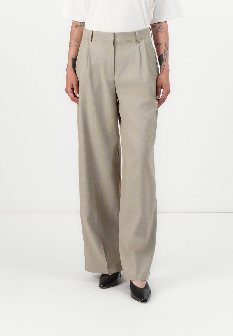 Beige wijde broek met plooien, gemaakt van een gladde stof. Draagt een witte top en zwarte puntige schoenen, met een getailleerde pasvorm.