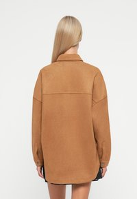 Brown oversized shirt jacket made of soft, textured fabric. Features a collar, dropped shoulders, and buttoned cuffs with a straight hem.