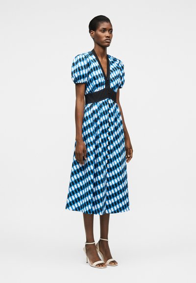 Γυναίκα στέκεται φορώντας midi φόρεμα με μπλε σχέδια, μαύρη ζώνη στη μέση και φουσκωτά κοντά μανίκια, συνδυασμένο με λευκά πέδιλα με ψηλό τακούνι.