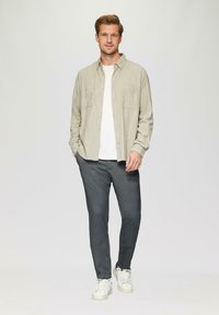 Chemise à manches longues gris clair avec deux poches poitrine, portée sur un t-shirt blanc ; associée à un pantalon texturé gris foncé et des sneakers blanches.